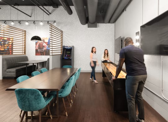 A community lounge with a game center and a table surrounded by eight blue chairs on hardwood-style flooring.