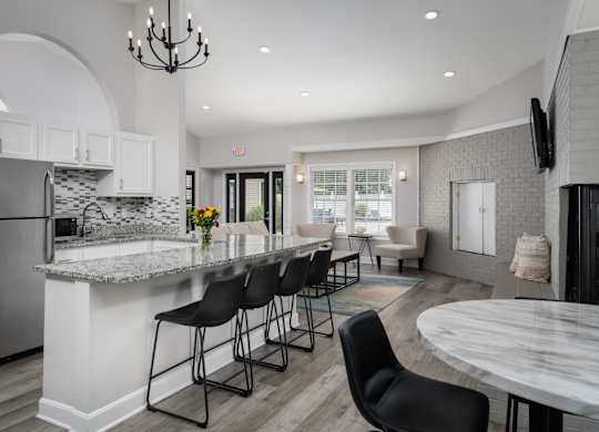 A kitchen in a clubhouse with stainless steel appliances and four barstools, next to a seating area with two chairs.