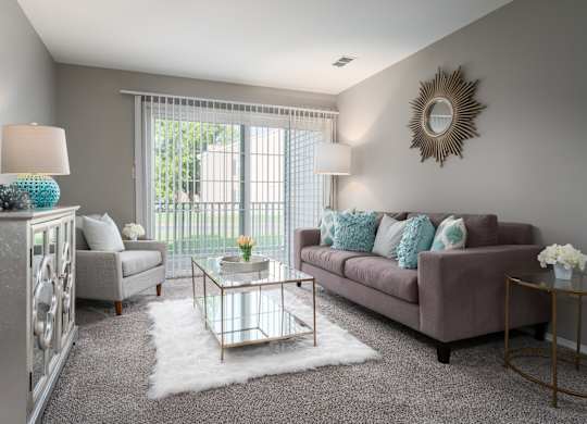 A furnished living room on carpeted flooring, next to a sliding glass door leading to a balcony.
