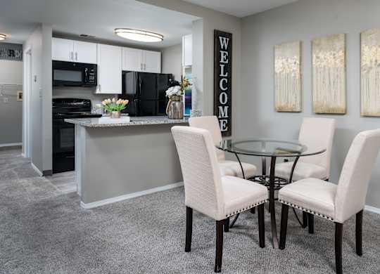 Dining room circular table surrounded by four chairs, situated next to a kitchen, on carpeted flooring.