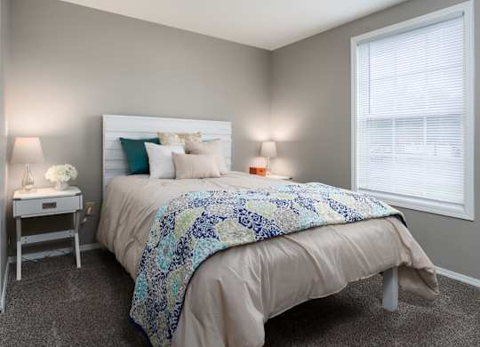 The Oaks bedroom with a bed, bedside tables, carpeted flooring, and a window for natural light.