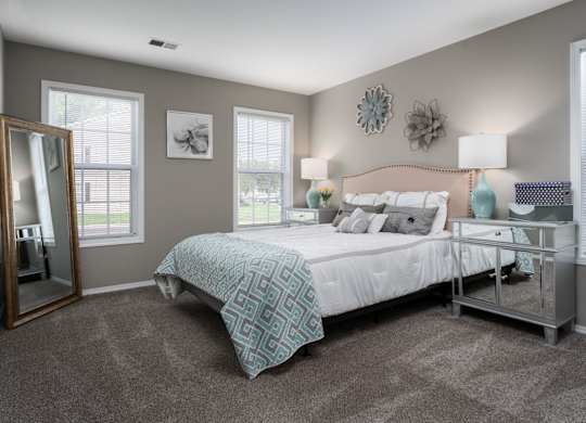 A bedroom with a bed and two bedside tables with a lamp on each side, carpeted floors, and two windows.