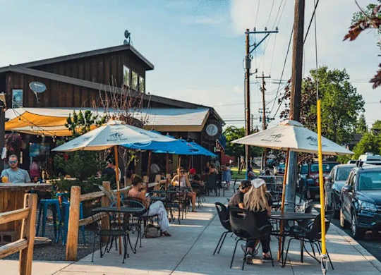 A group of people are sitting at tables under umbrellas outside a wooden building.