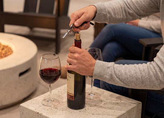 A person is opening a bottle of wine in front of a fire pit.