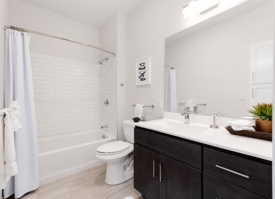 Model apartment bathroom with large mirror, white counter and black cabinet.