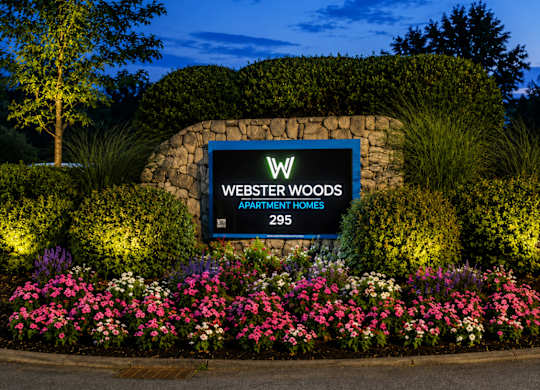 A sign for Webster Woods Apartment Homes is surrounded by flowers and greenery.