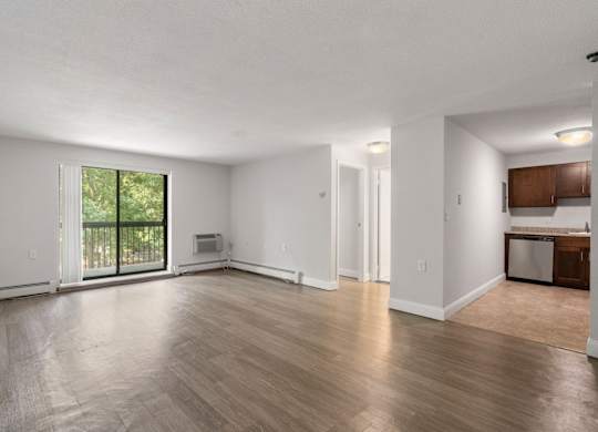 A spacious living room with wooden flooring and a sliding glass door leading to a balcony.