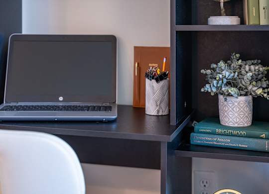 A laptop is on a desk with a white chair, a pencil holder, and a book titled "Brightwood" on a shelf at the Mastlight.