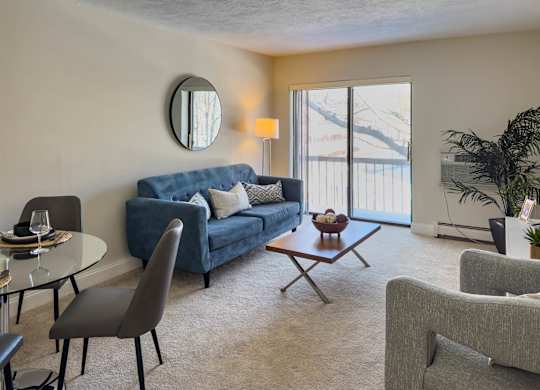 A living room with a blue couch, grey armchairs, a glass table, and a round mirror on the wall.