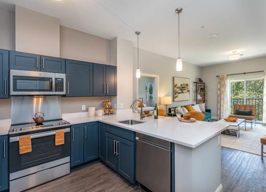 The Residences at Crosspoint open-plan kitchen and living area includes navy blue shaker cabinets, stainless steel appliances, a white island countertop with stools, and seating near a sliding glass door leading to a balcony.