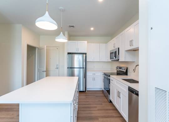 Bright, modern kitchen with a long white island, stainless-steel appliances and pendant lighting beneath white Shaker cabinets.
