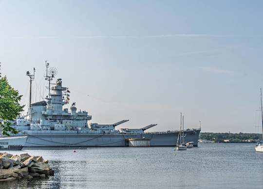 Historic battleship anchored on a calm river alongside small sailboats and tree-lined shoreline.