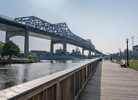 Boardwalk beneath a steel truss bridge, looking toward moored naval ships along the sparkling river.