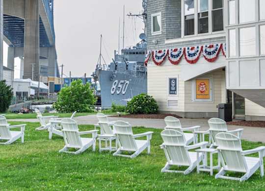 Lawn with white Adirondack chairs beside a carousel pavilion draped in patriotic bunting, with destroyer 850 and bridge backdrop at Battleship Cove