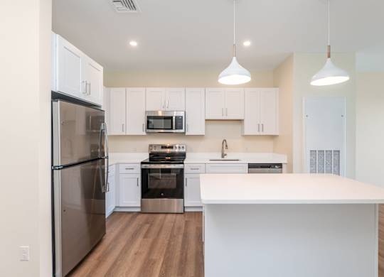 Open kitchen lined with white Shaker cabinets, quartz island workspace and twin pendant lights, plus stainless fridge and range.