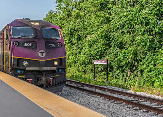 Purple MBTA commuter-rail locomotive arriving at Fall River Depot, framed by tree-lined tracks and platform edge.