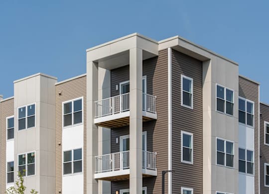 Detail of building façade showing stacked private balconies, taupe siding and vertical columns against a clear blue sky.