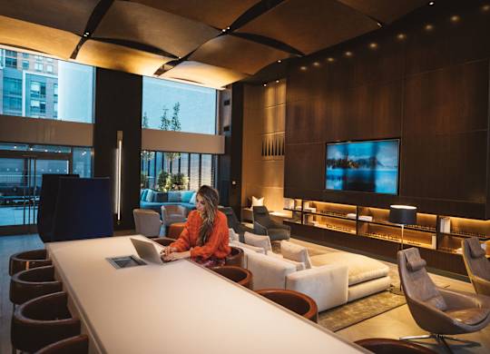 A woman in an orange shirt is sitting at a long white table in a room with a large screen on the wall.