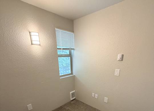 A room with a carpeted floor, a window with blinds, and a wall-mounted light fixture.