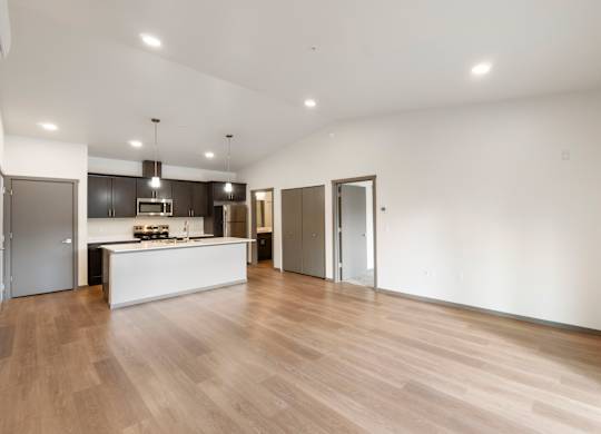 A spacious room with wooden flooring and a kitchen area in the background.