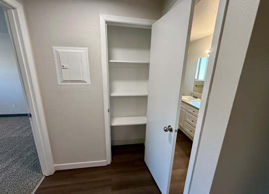 A white door is open to reveal a small pantry with a shelf and a light switch on the wall.