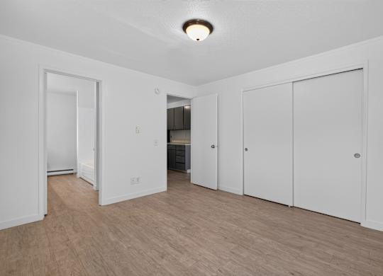 A room with white walls and wooden flooring with a refrigerator and cupboards in the background.