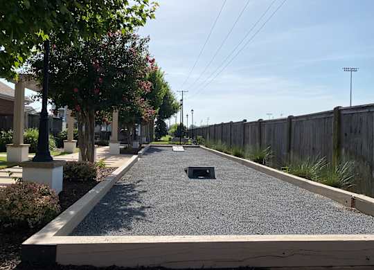 A long, narrow, gravel-covered pathway stretches into the distance between a wooden fence and a row of trees.
