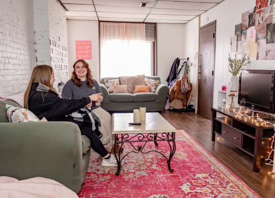 two women sit in a living room with a red rug and a television at University Commons Apartments, Oakland, Pittsburgh