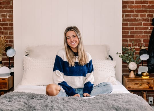 a woman sitting on a bed with a brick wall behind her at University Commons Apartments, Oakland, Pittsburgh