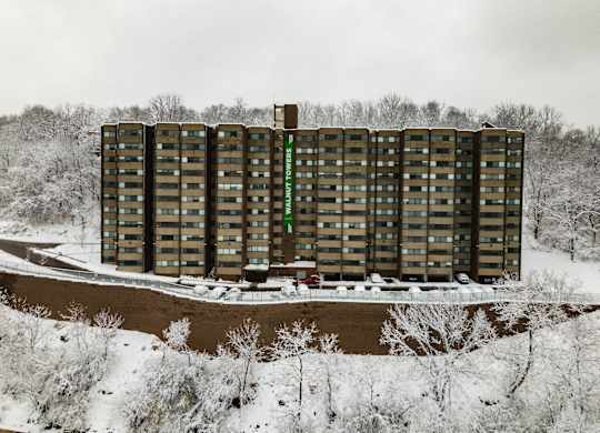 A tall building with a green sign on the side stands in front of a snowy landscape at Walnut Towers at Frick Park, Pittsburgh, PA