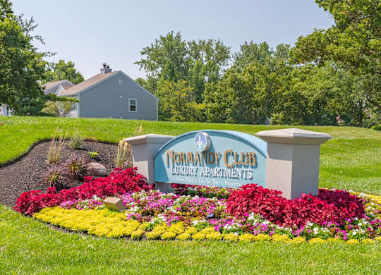 A sign for Normandy Club Luxury Apartments is surrounded by flowers.