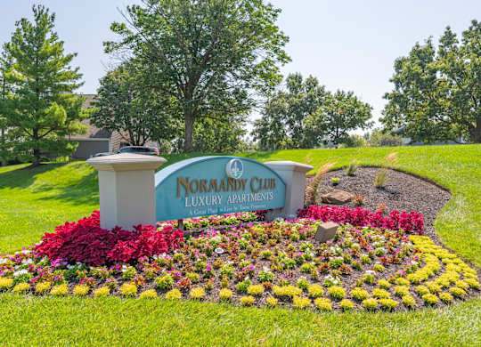 A sign for the Normandy Club Luxury Apartments is surrounded by flowers.