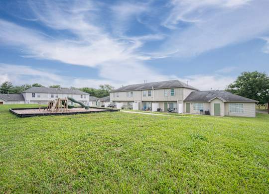 A large grassy field with a playground and houses in the background.