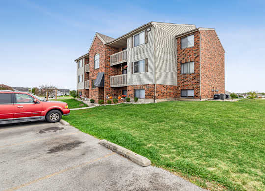 A red car is parked in a parking lot in front of a brick apartment building.