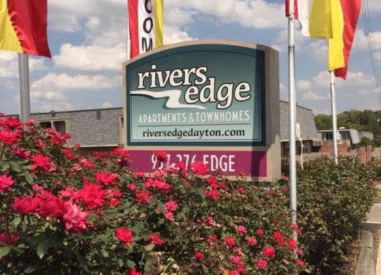 a sign for rivers edge apartments and townhomes with flags and flowers at Rivers Edge Apartments and Townhomes, Dayton, OH