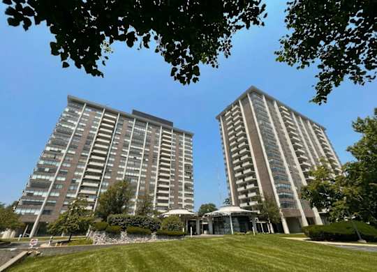 Two tall buildings with balconies are surrounded by a grassy area and trees.