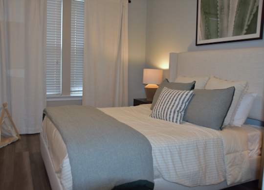 A bed with a grey blanket and pillows is in the foreground of a room with a lamp and a painting on the wall.