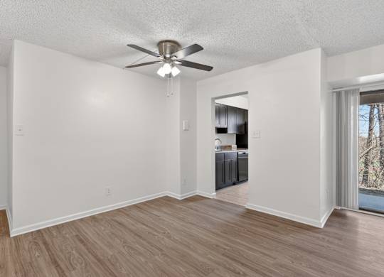 A room with a ceiling fan and light fixture, hardwood floors, and a kitchenette in the background.