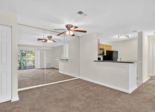 A spacious living room with a ceiling fan and a kitchenette in the background.