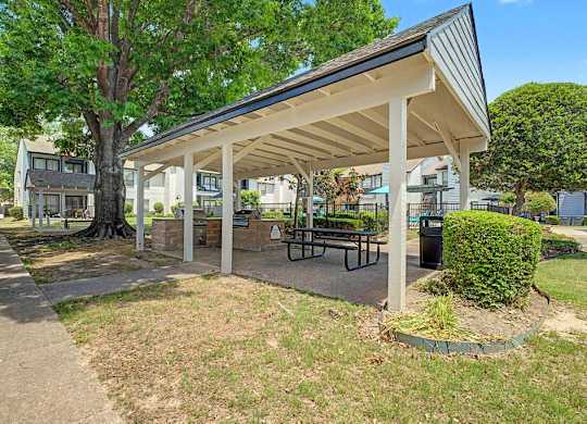 A covered picnic area with benches and a table is surrounded by greenery.