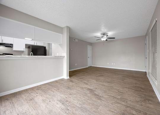 A room with a ceiling fan and light, a kitchenette with white cabinets and a black fridge, and a wooden floor.