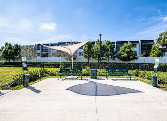 A playground with a round platform and a shade structure is surrounded by green bushes and trees.