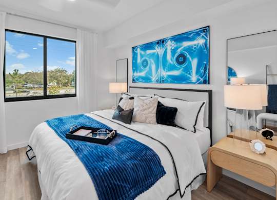 A bedroom with a large bed, a blue and white comforter, a wooden nightstand, and a large blue abstract painting on the wall.