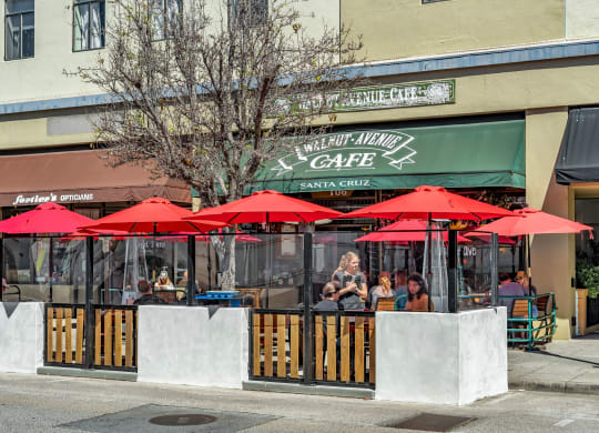 the exterior of a restaurant with red umbrellas and people sitting at tables