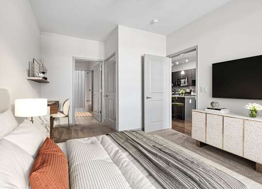 Bedroom at 7001 Arlington at Bethesda in Bethesda, MD, featuring a bed, wall-mounted TV, writing desk, carpeted flooring, and doorways to the bathroom and kitchen.