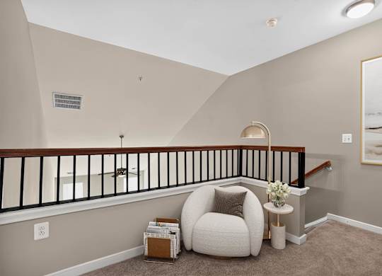 Virtually staged open loft at Enclave at Emerson overlooking the main living area, with neutral carpeting, a rounded lounge chair, side table and floor lamp.