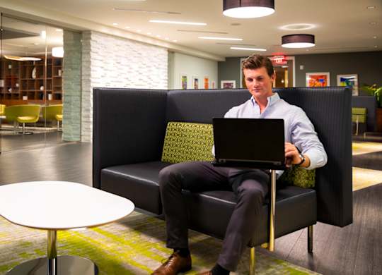 A man in a white shirt and black pants is sitting on a black couch with a laptop on his lap.