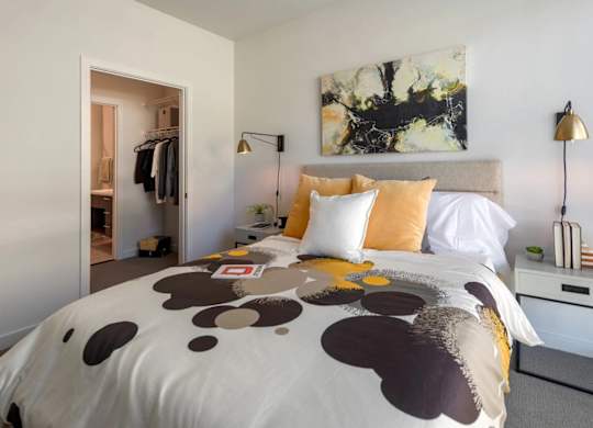A bed with a white comforter and a brown and black pattern is in the foreground of a room with a painting on the wall and a clothing rack in the background.