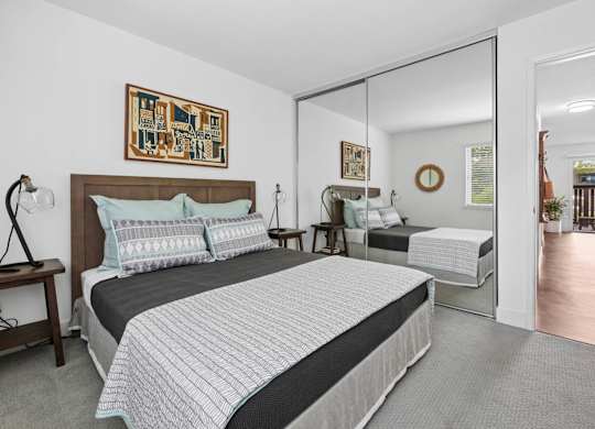 Bedroom with wood bed frame, nightstand, lamp, and window with blinds at Old Orchard Apartments