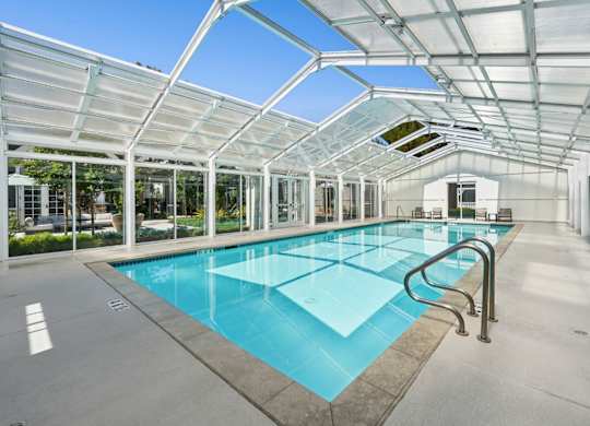 Indoor swimming pool at Pescadero Apartments with glass enclosure, lounge seating, and natural light.